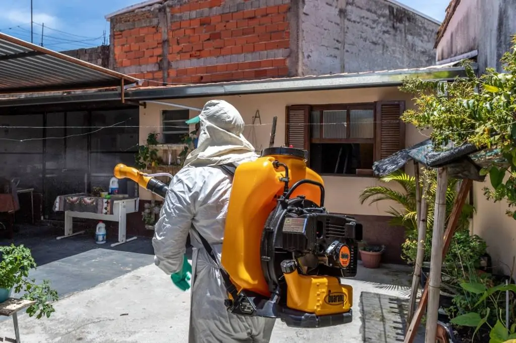 Homem veste roupas e usa aparelho para espalhar inseticida dentro de residências. Crédito: Divulgação/PMG.