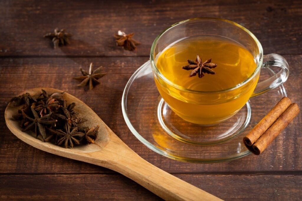 Xícara e pires de vidro com chá de anis-estrelado e canela. Colher de pau ao lado com anises-estrelados