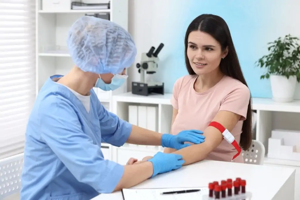 Profissional de saúde usando jaleco azul e máscara enquanto faz coleta de sangue em paciente mulher de cabelos longos e escuros