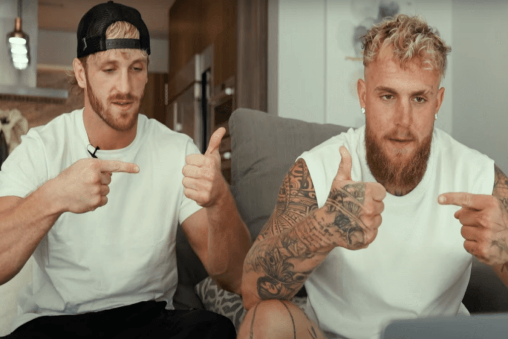 Imagem de dois homens vendo e imitando vídeo, os dois estão usando camisetas brancas e um deles usa um boné preto