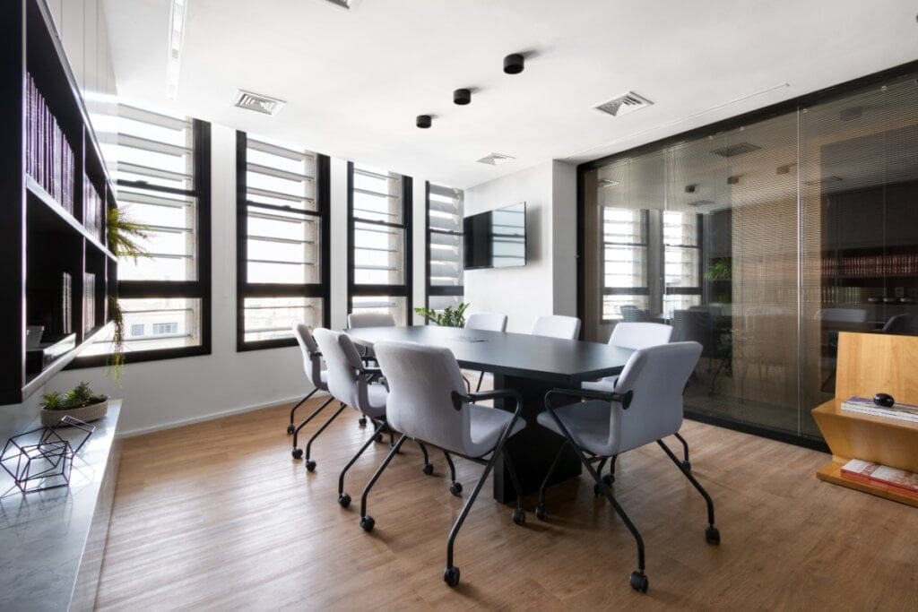 Sala moderna com piso amadeirado, janelas com brises verticais pretos e cadeiras de escritório ao redor da mesa