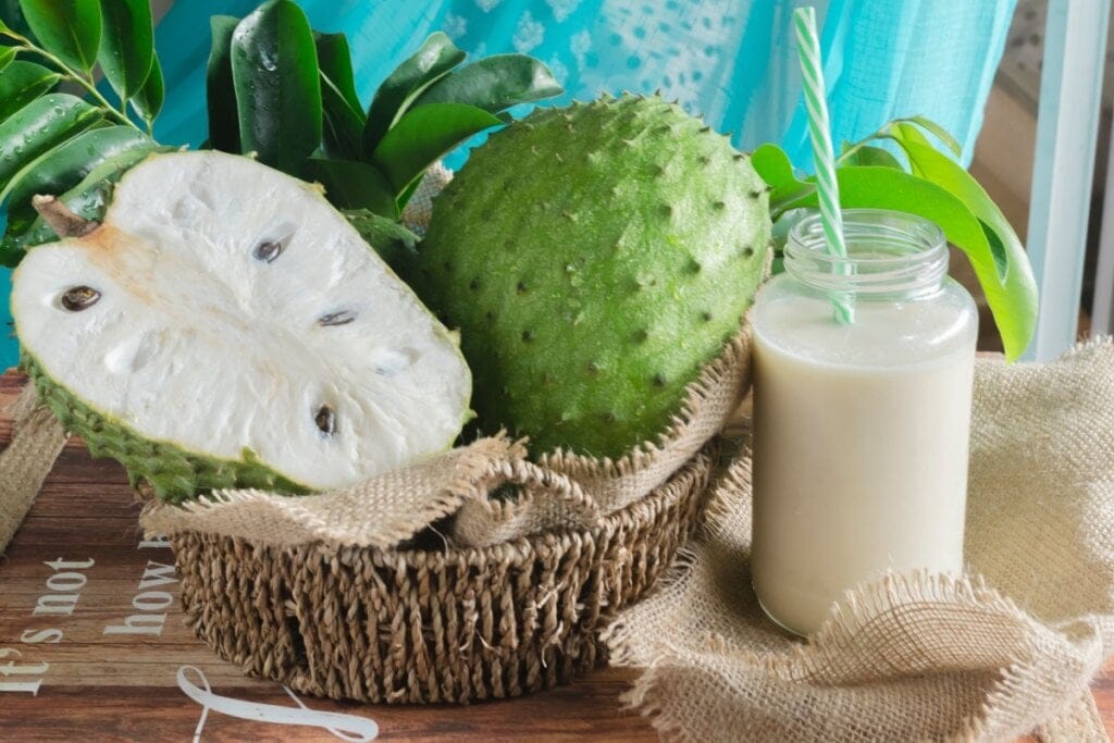 Uma graviola inteira e uma metade dentro de cesto de palha e um copo de suco de graviola ao lado