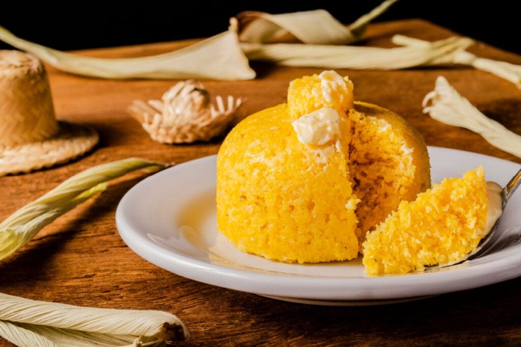 Cuscuz com manteiga em prato branco. Ao fundo palhas de milho