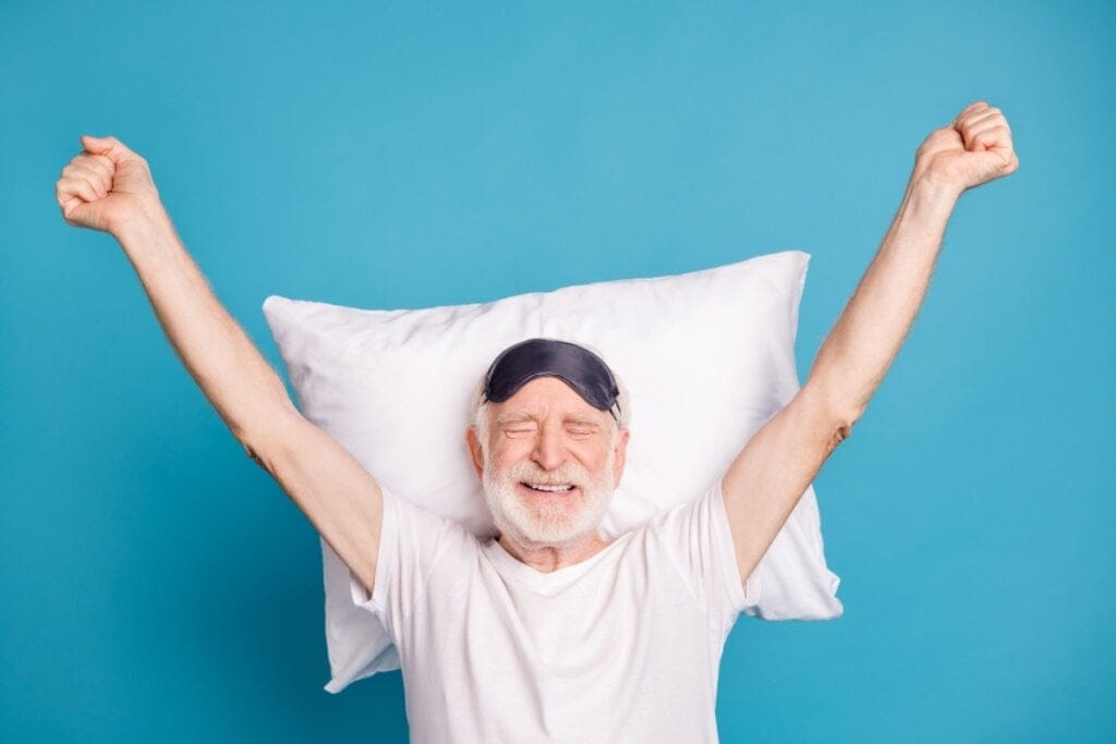 Homem idoso usando camiseta branca e máscara de dormir preta encostado em travesseiro branco em fundo azul 