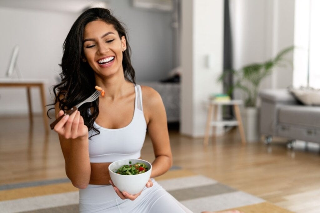 Mulher comendo salada sentada na sala, ela está vestindo roupas de academia na cor branca
