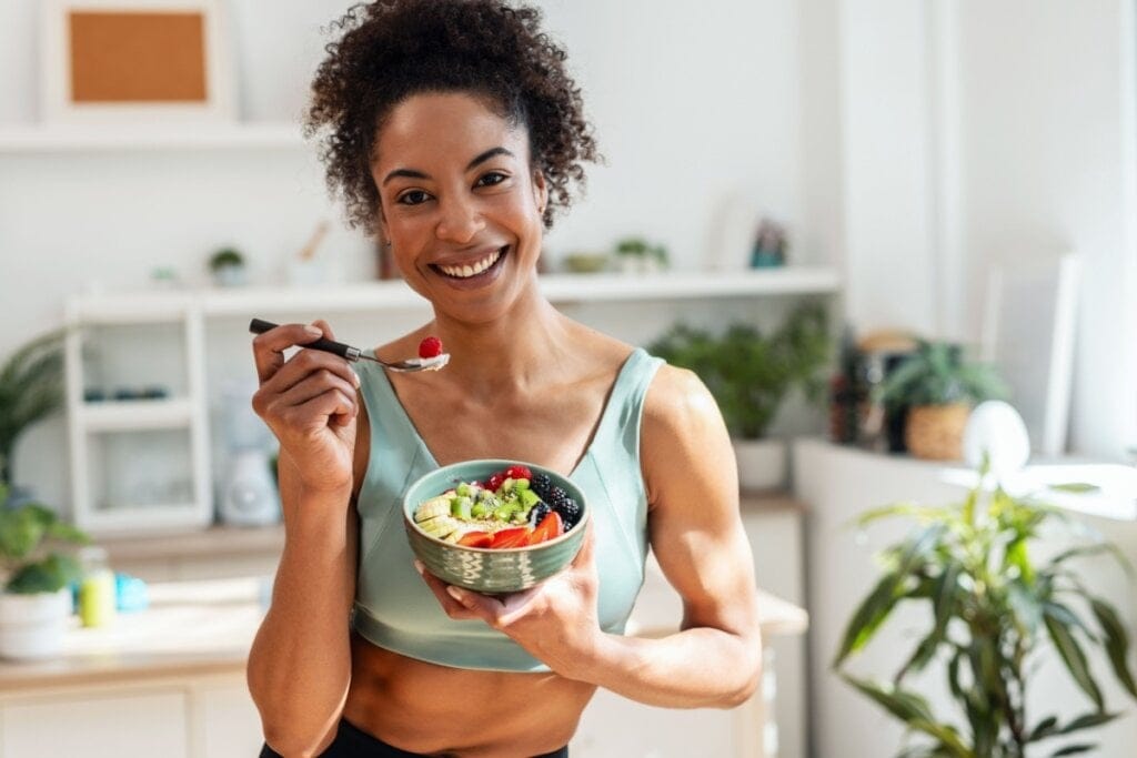Mulher negra com cabelo cacheado preso em rabo de cavalo e usando top de academia verde com uma tigela de frutas na mão e um garfo em outra