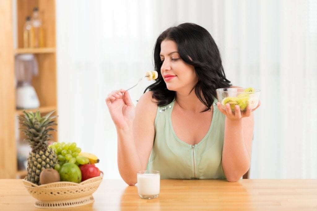 Mulher sentada à mesa segurando recipiente de frutas e uma colher, prestes a comer, com um copo de iogurte natural e uma cesta de frutas à frente