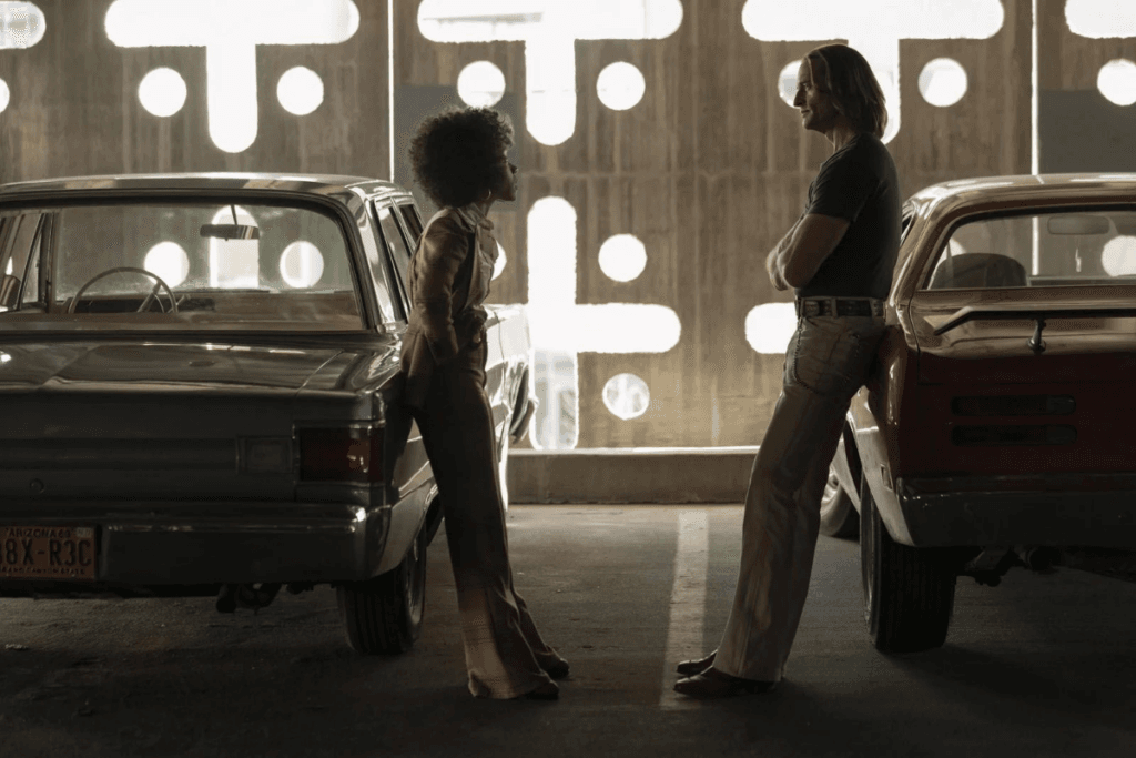 Uma mulher e um homem usando roupas dos anos 70 encostados em carros da época