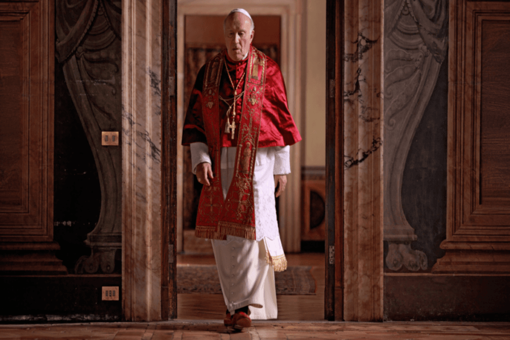 Homem vestido com roupas litúrgicas de Papa em vermelho e branco saindo de uma sala
