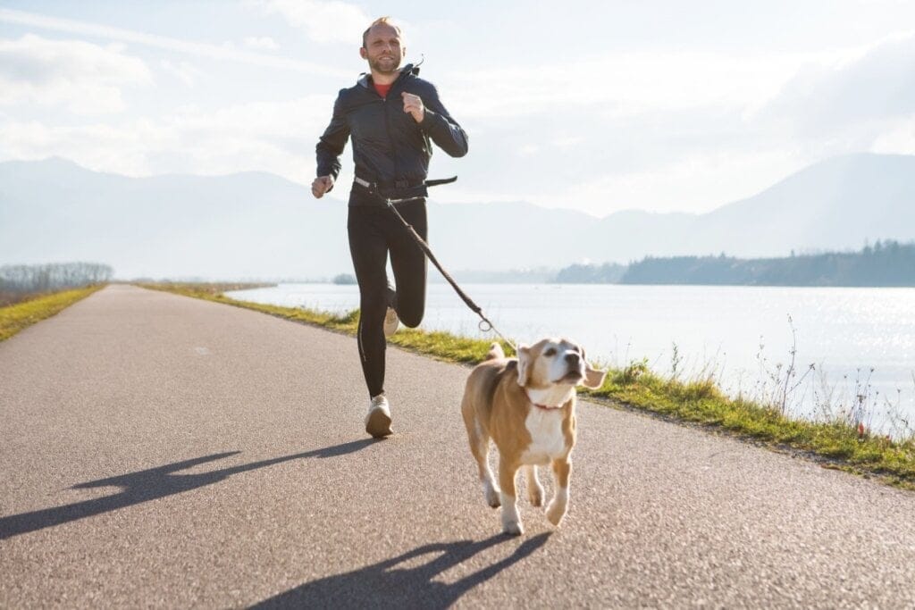 Homem vestindo legging e blusa de frio praticando canicross com um cachorro em frente a um lago