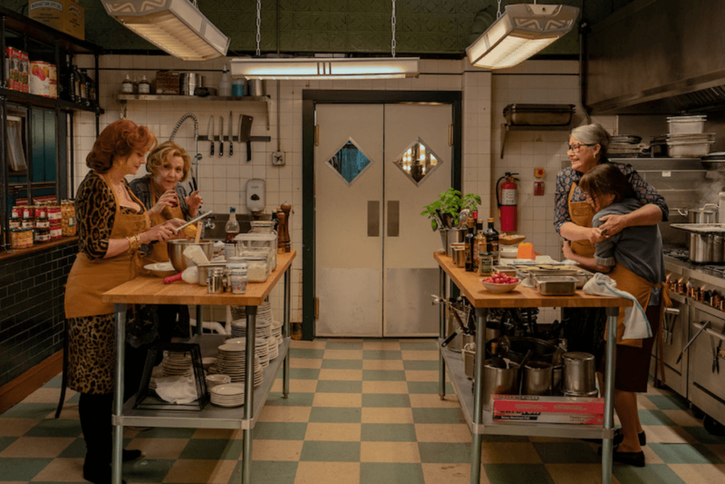 Uma cozinha com duas bancadas de madeira e com 4 mulheres cozinhando, cada uma está usando um avental amarelo