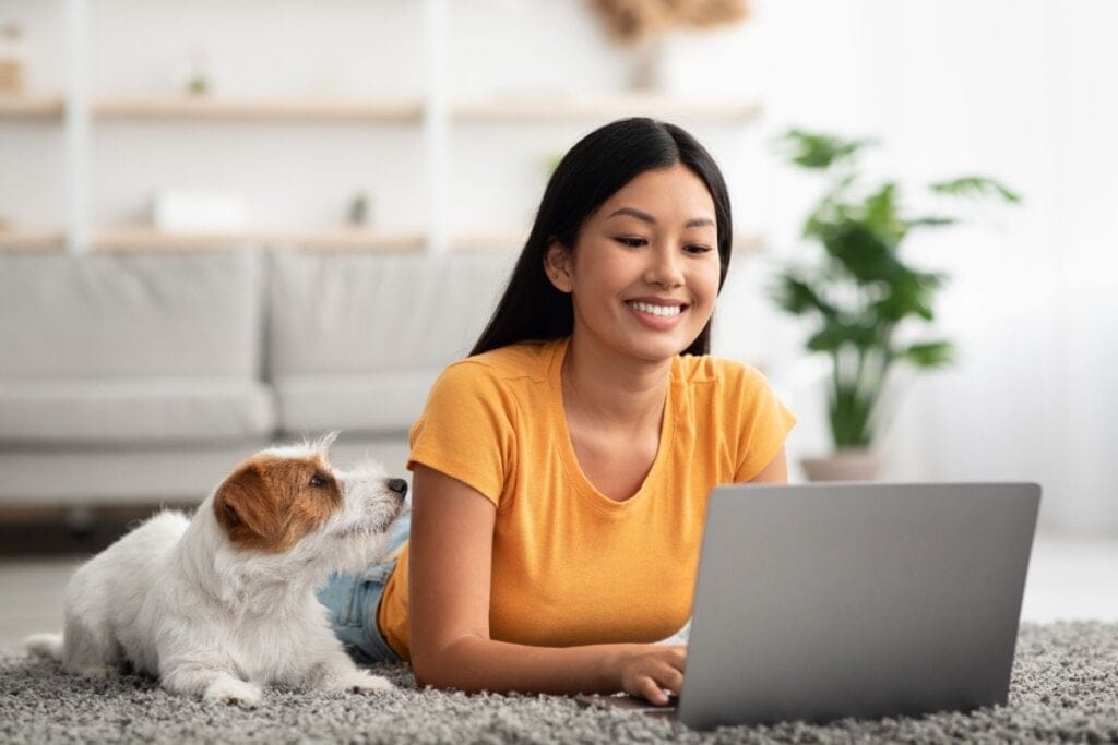 Imagem de uma mulher deitada no chão teclando no notebook com um cachorro ao lado dela