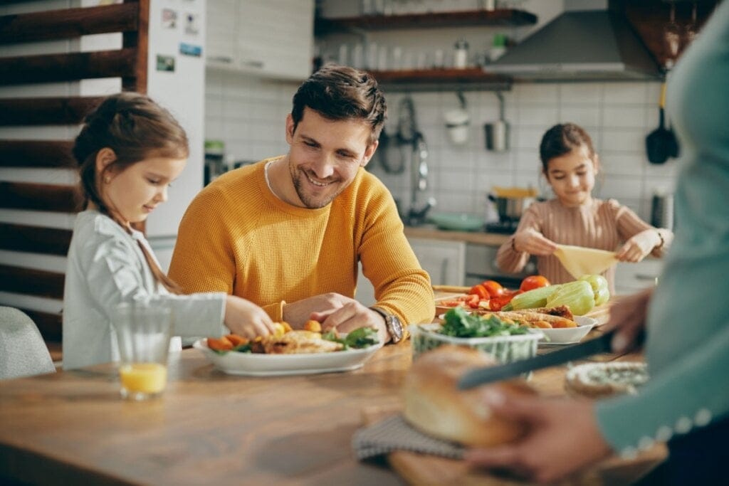 Duas meninas, ao lado de um homem, em uma cozinha, comendo vegetais