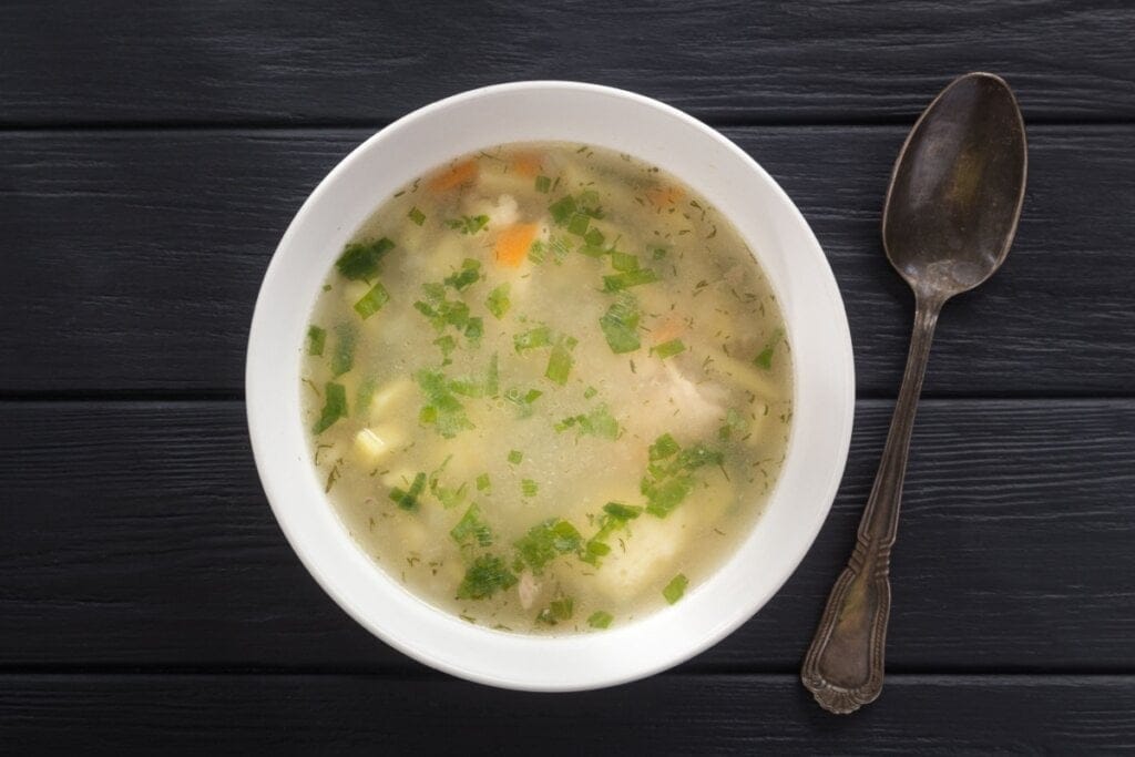 Caldo de frango servido em tigela de cerâmica branca, com colher ao lado em cima de mesa de madeira