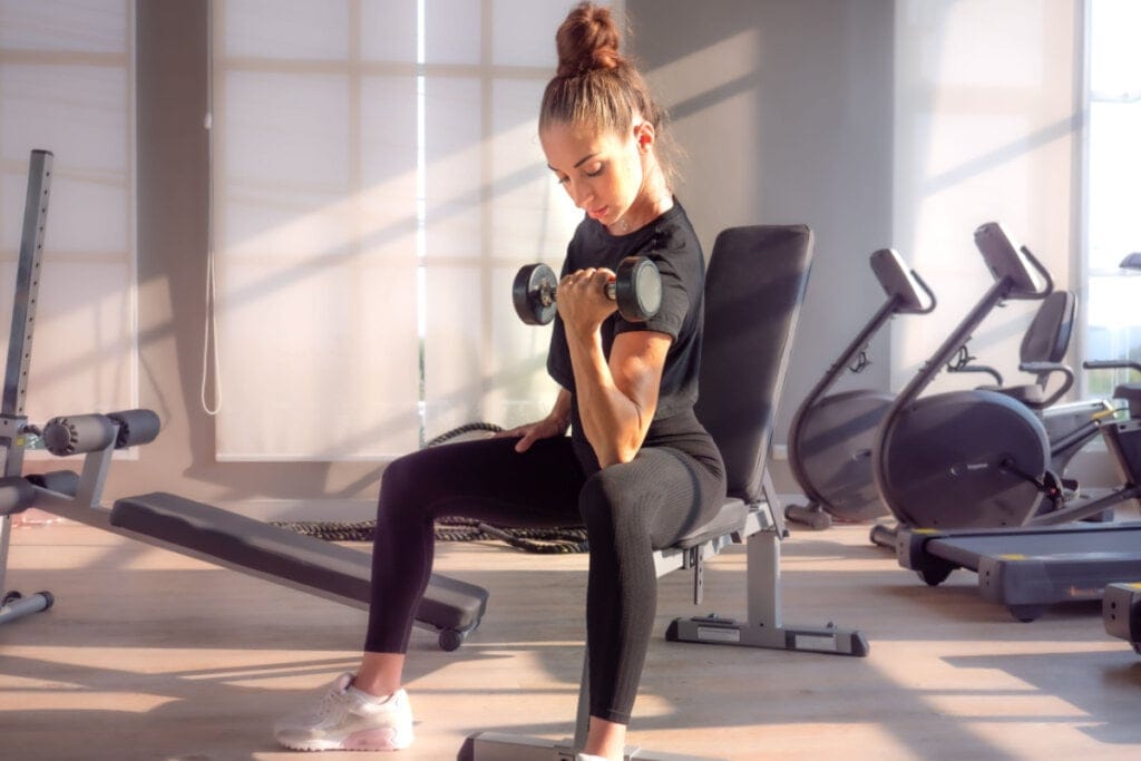 Mulher com roupa preta sentada na academia levantando peso no braço