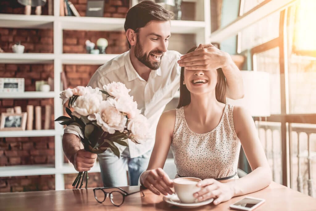Homem segurando um buquê de rosas com a mão nos olhos de uma mulher sorrindo