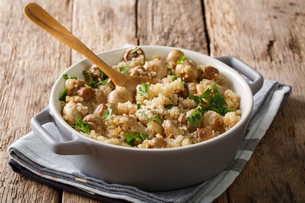 Panela branca com quinoa, cogumelos e salsinha em cima de uma mesa de madeira