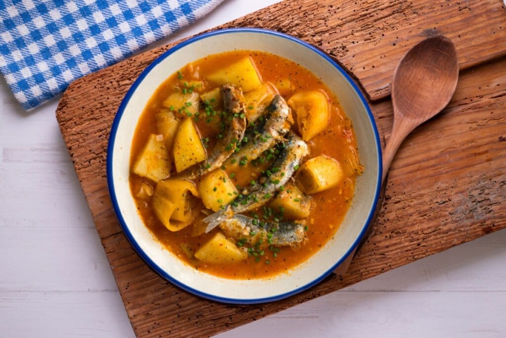 Prato fundo com ensopado de sardinha fresca, batatas em cubos e rodelas de alho-poró em caldo avermelhado, decorado com cebolinha