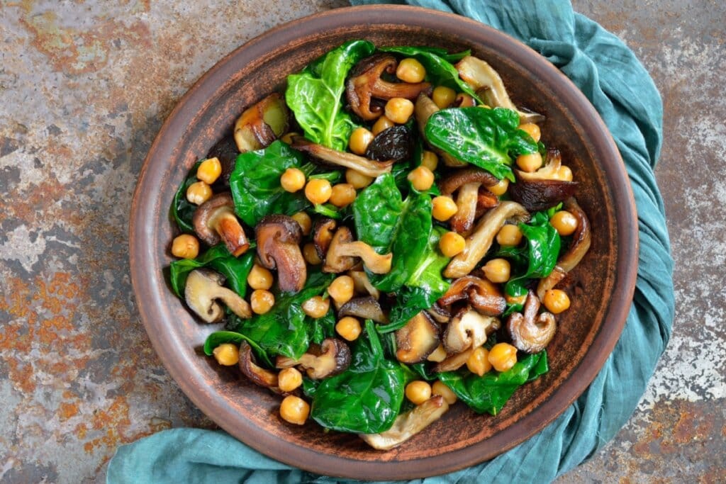 Salada de espinafre, grão-de-bico e cogumelos em recipiente marrom com pano azul-esverdeado ao lado