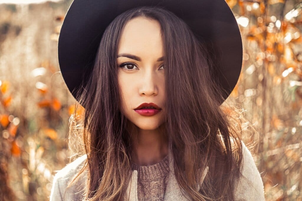 Mulher usando um chapéu preto e um batom vermelho, posando para foto em uma paisagem com folhas secas 