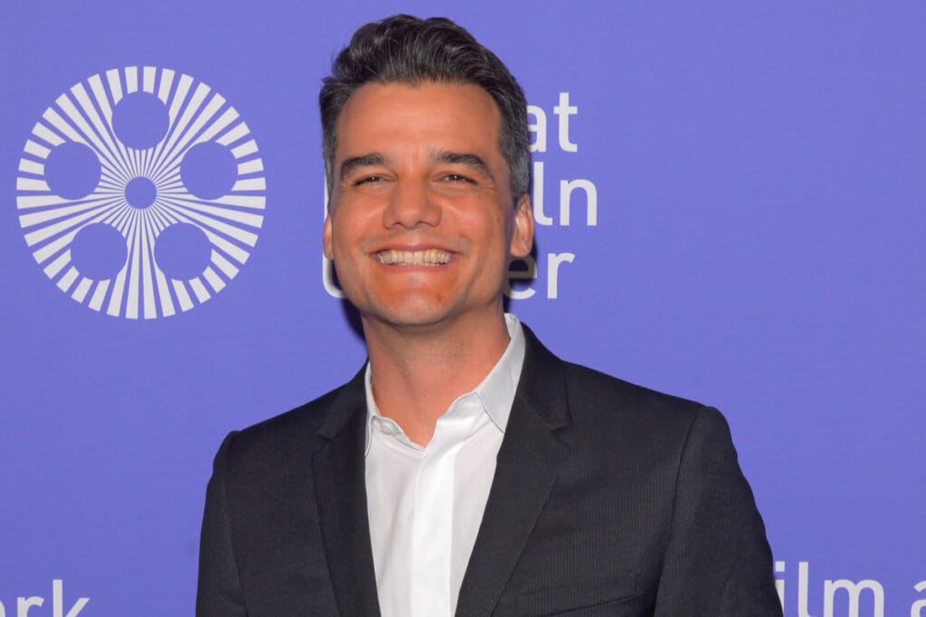 Homem com cabelo preto curto, usando camisa branca e terno preto sorrindo posando para foto em fundo roxo