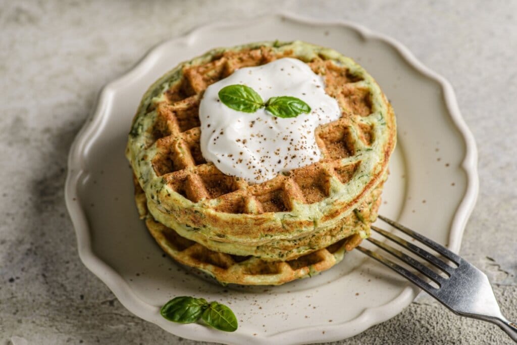 Waffle de abobrinha servido com molho de iogurte em prato de cerâmica branco em cima de mesa de pedra 