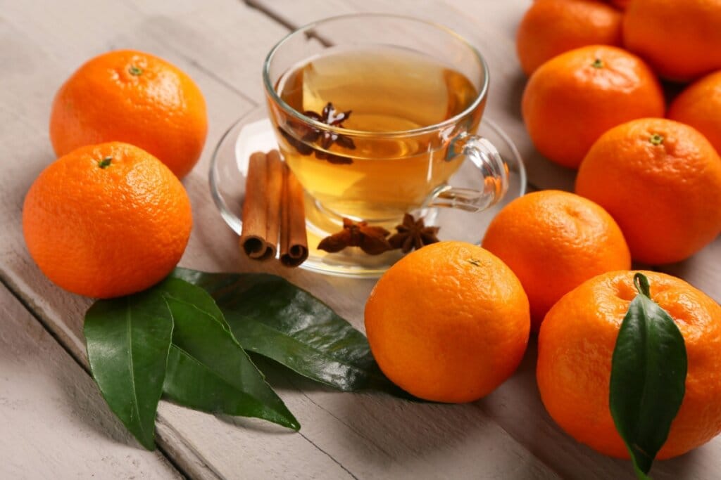 Xícara de vidro com chá de tangerina com anis-estrelado e canela em cima de uma mesa de madeira com tangerinas
