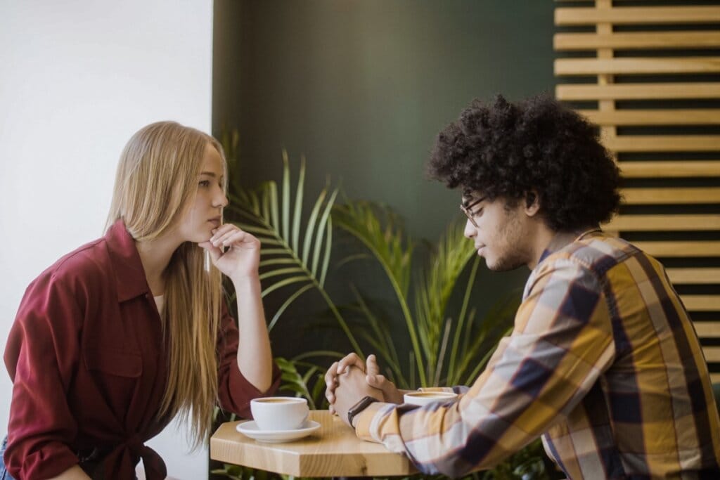 Rapaz de cabelos escuros e cacheados em mesa com garota loira séria