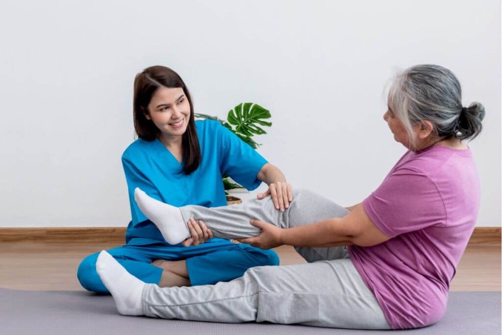 Médica com o cabelo liso solto, usando um uniforme azul auxiliando a fisioterapia no joelho de uma paciente idosa usando uma camiseta roxa, uma calça cinza e meia branca