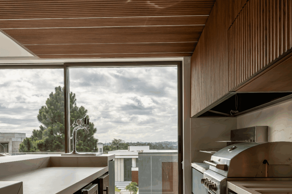 Área gourmet com churrasqueira e forro em madeira e ármarios em madeira e bancada de mármore com parede de vidro ao fundo