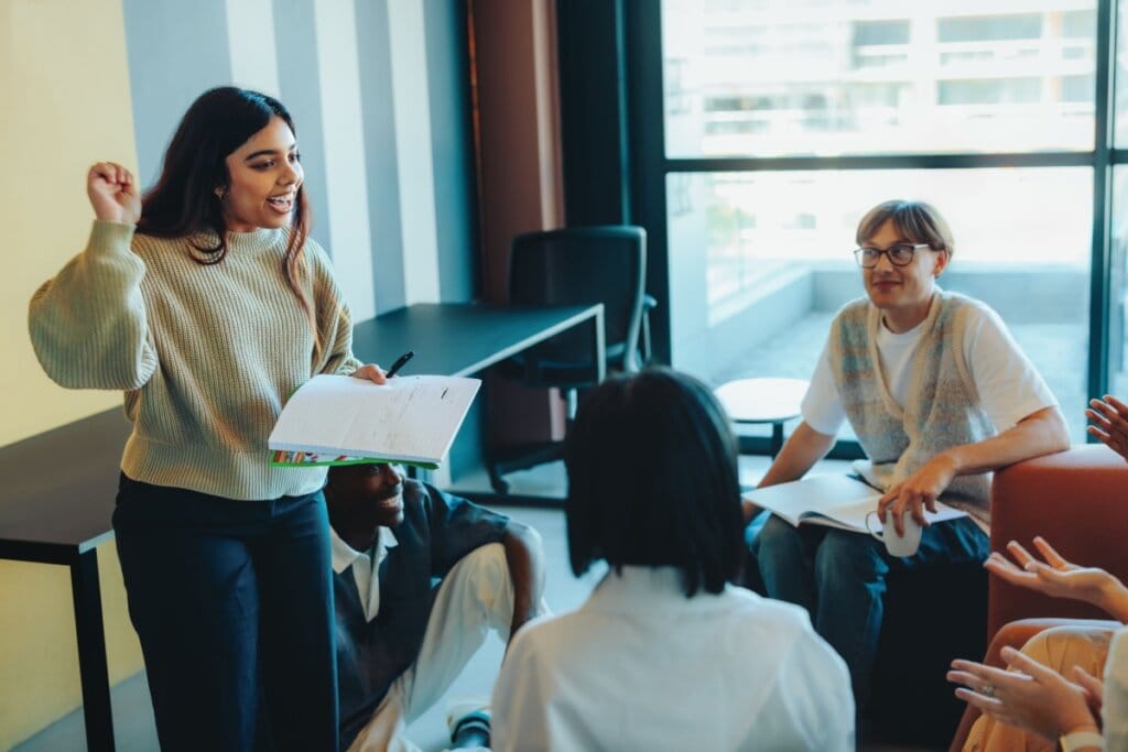 Grupo de estudantes reunidos e conversando, uma delas está de pé com um caderno na mão explicando algo 
