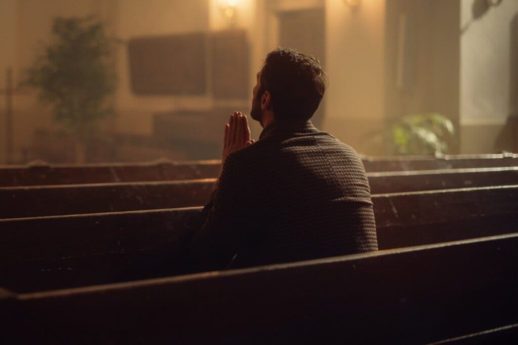 Homem usando camisa marrom orando sentado em banco de igreja