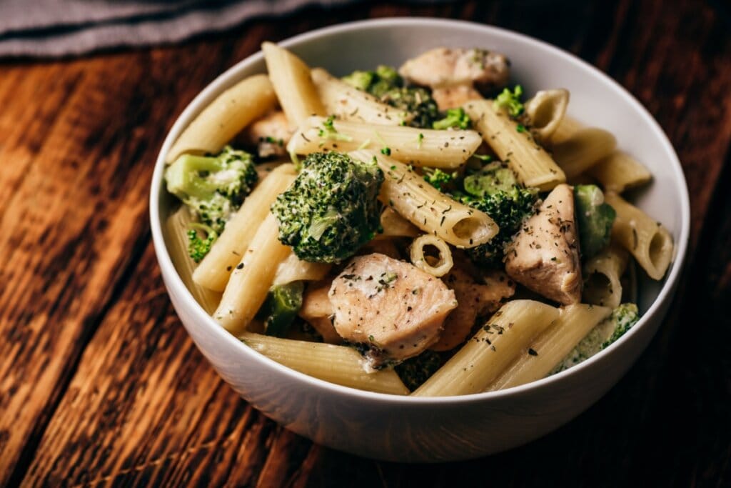Macarrão integral com frango e brócolis servido em tigela branca de cerâmica em cima de mesa de madeira