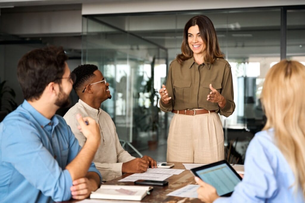 Mulher conversando com equipe de trabalho; ela tem o cabelo curto castanho e está usando uma camisa de botões verde e calça bege