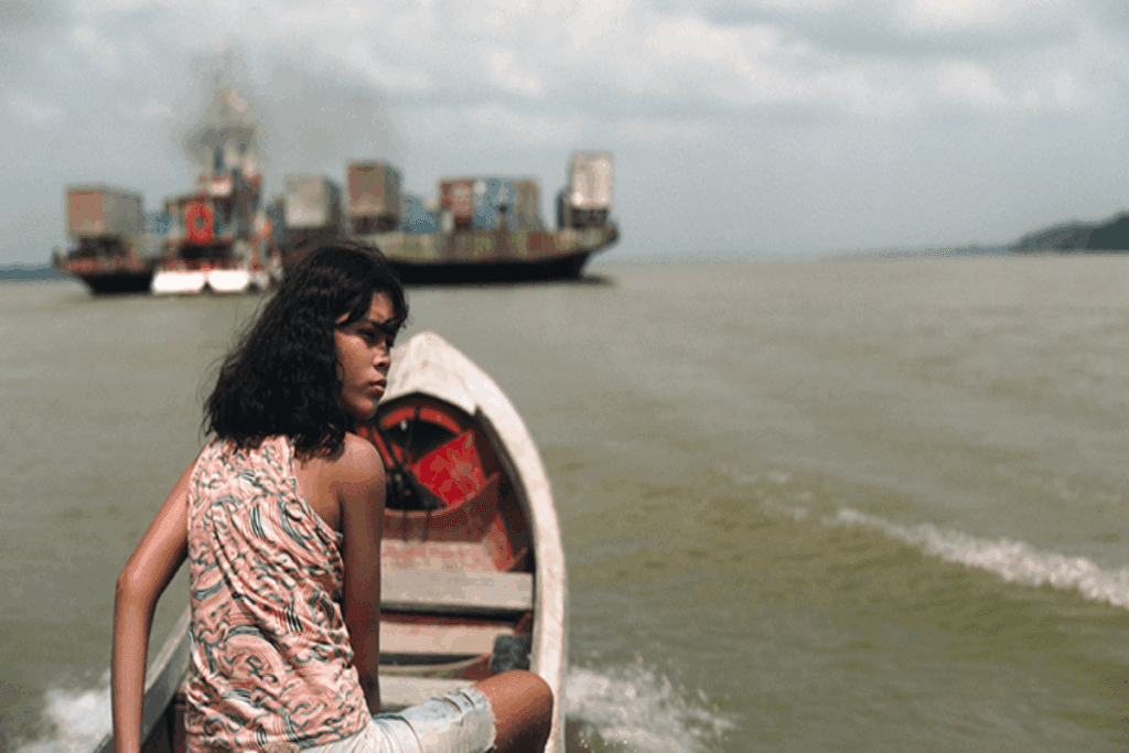 Uma jovem com cabelo curto, usando uma regata estampada e bermuda jeans andando em barco com barcos ao fundo