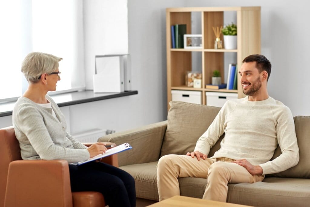 Homem com cabelo curto, de barba e usando camisa de manga longa e calça bege, sentado em sofá bege conversando com psicóloga que tem o cabelo curto, grisalho e está usando um suéter cinza, calça preta e está sentada em uma cadeira marrom