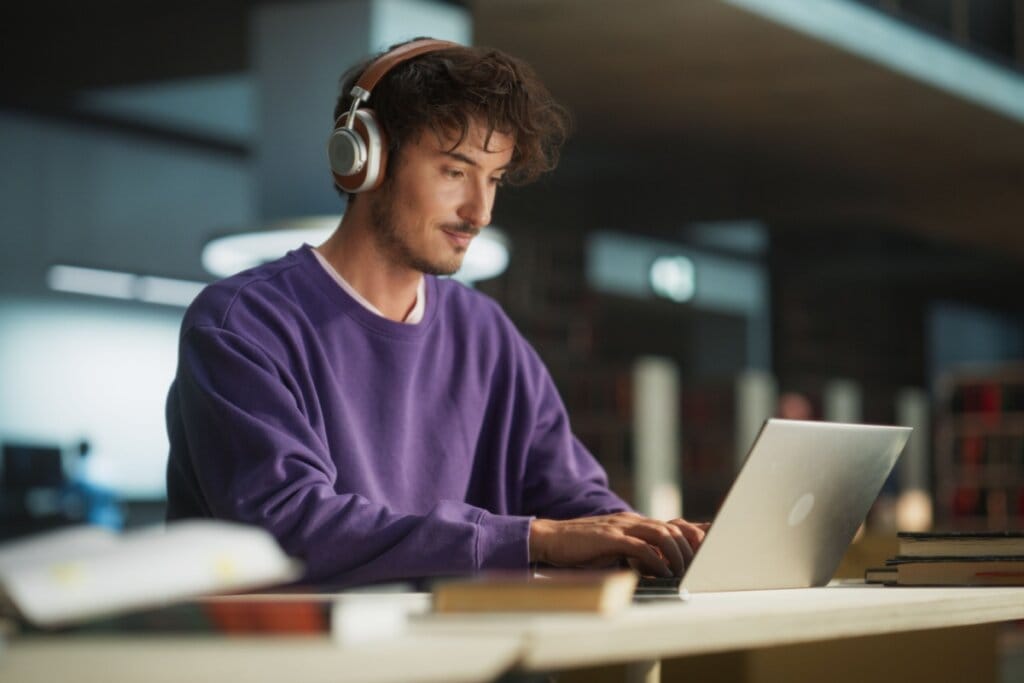 Jovem com o cabelo curto e usando blusa de moletom roxa, e fones de ouvido prata,  estudando no notebook