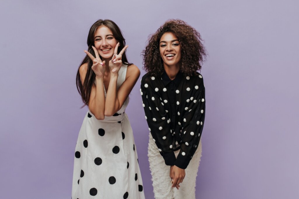Imagem de duas jovens, uma tem o cabelo liso e está solto, e ela está usando um vestido branco com bolinhas pretas e a outra tem o cabelo curto cacheado e está usando uma calça branca e uma blusa de frio preta com bolinhas brancas em fundo roxo