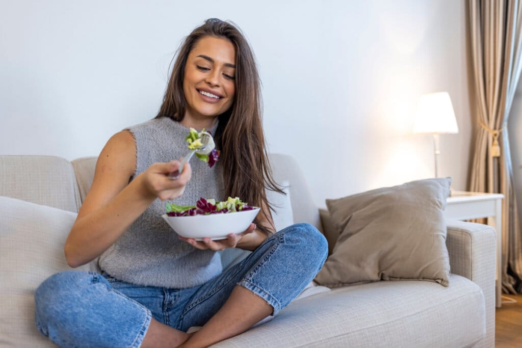 Mulher sentada no sofá comendo salada em pote branco