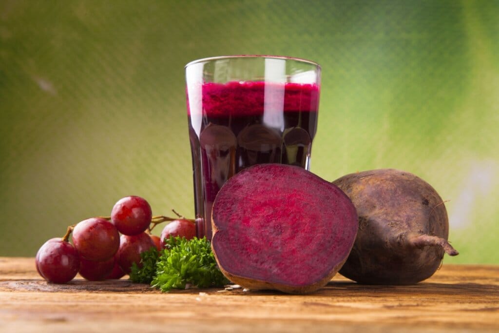 Copo de vidro com suco de beterraba ao lado de um cacho de uva e duas beterrabas 