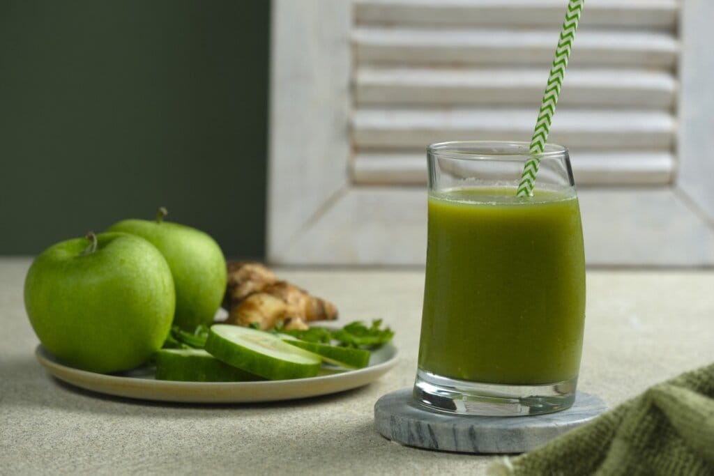 Copo de vidro com suco verde e ao lado de um prato com maçã-verde, rodelas de pepino e gengibre 