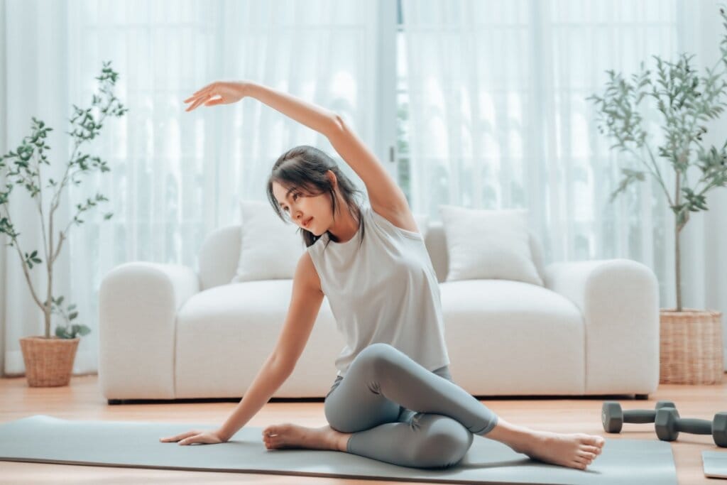 Mulher sentada em tapete realizando alongamento lateral em casa com halteres ao lado
