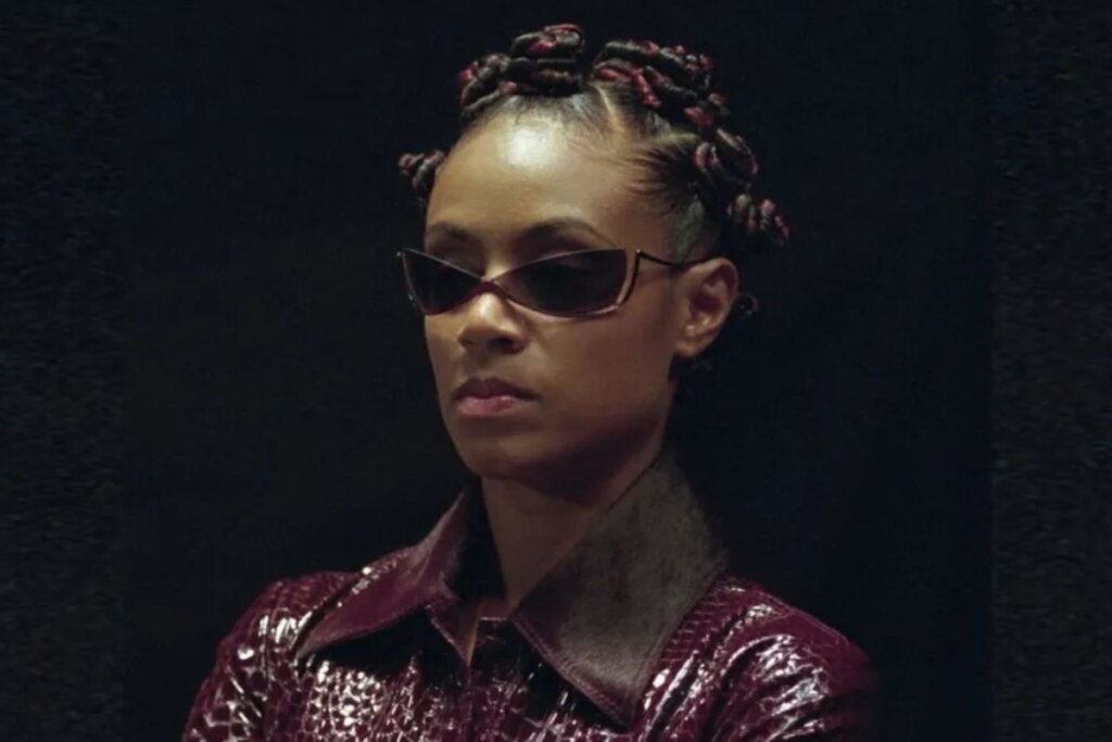 Jada Pinkett Smith em Matrix, usando roupa de couro vermelha, óculos de sol e pequenos coques no cabelo 