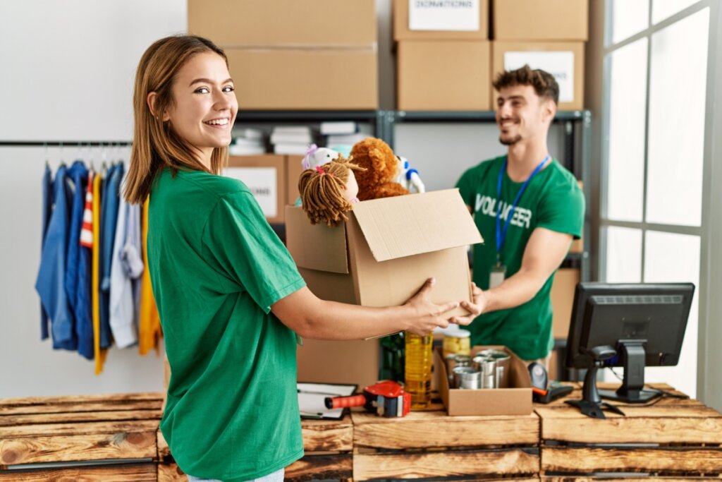 Dois voluntários, um homem com cabelo curto e barba e uma mulher com cabelo curto liso, ambos usando camisetas verdes, organizando caixas de doação