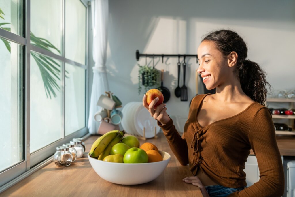 Jovem com cabelo preso em rabo de cavalo usando blusa de manga longa marrom escolhendo fruta na fruteira
