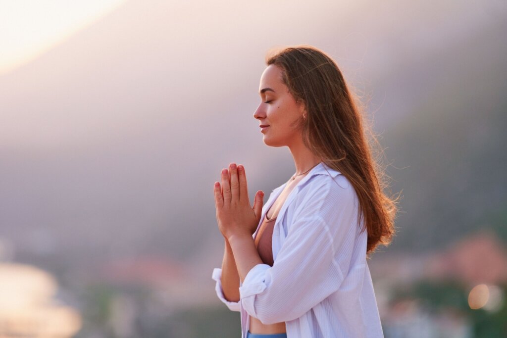 Mulher com o cabelo castanho solto, usando camisa de botões brancas e top marrom com as mãos juntas meditando ao ar livre 