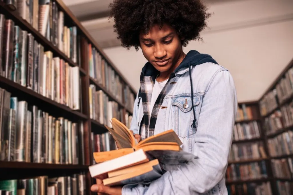 Jovem em uma biblioteca, segurando uma pilha de livros e lendo um deles