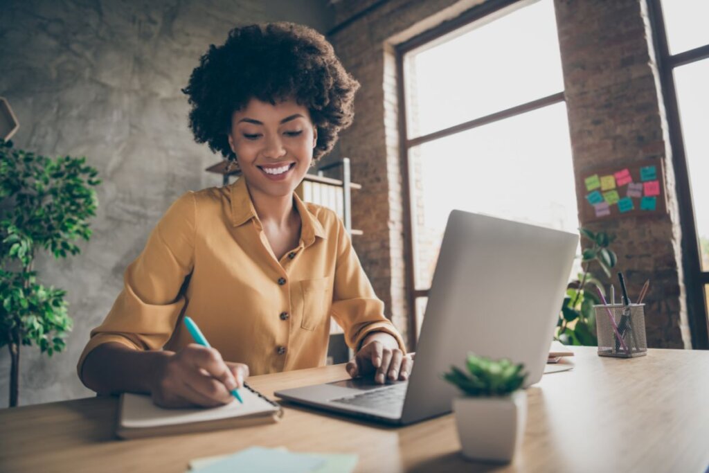 Mulher sentada com computador aberto e escrevendo em um caderno