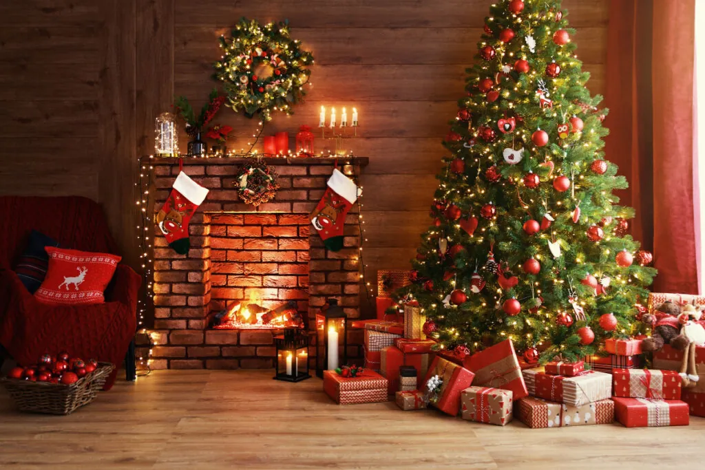 árvore de natal com enfeites vermelhos com presentes com embalagens vermelhas em volta. lareira ao lado com guirlanda, pisca-pisca e meias de natal