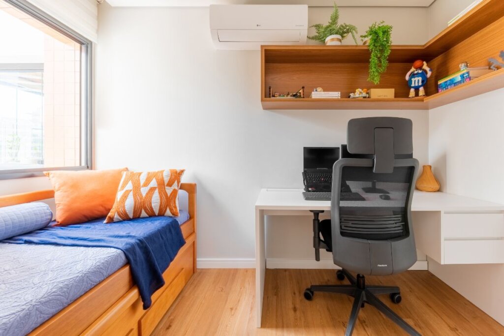Quarto com cama de madeira forrada com lençol branco e, ao lado, uma mesa branca com computador e cadeira preta a frente